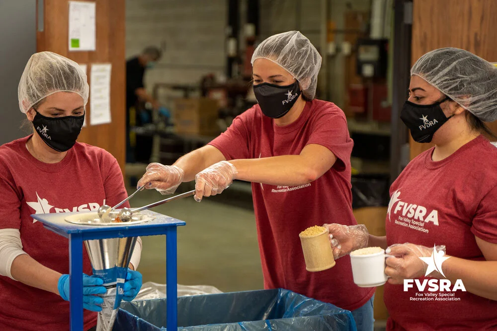 Three FVSRA staff members in hairnets and gloves packing meals at a Feed My Starving Children facility