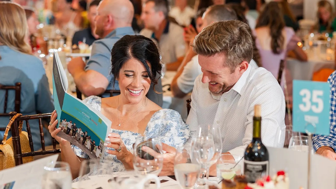 Two guests smiling together at a table while reading the event program at the Outreach gala