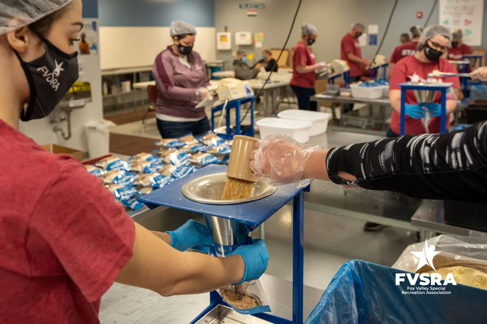 An FVSRA staff member funneling rice into a meal bag while another volunteer adds ingredients