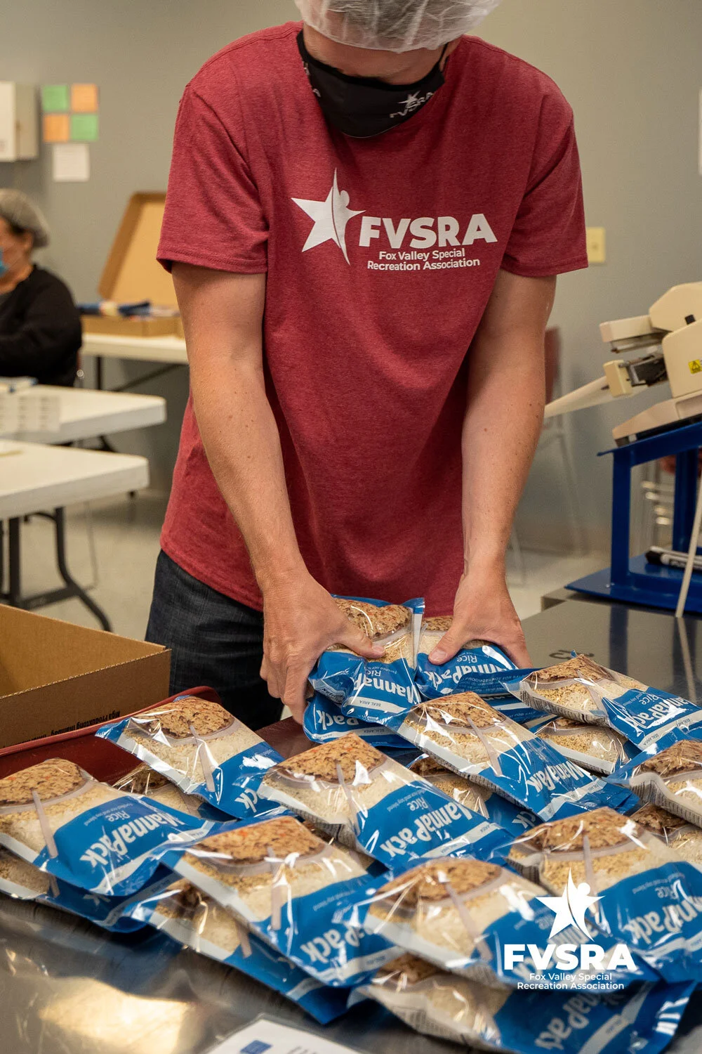 An FVSRA staff member in a hairnet placing sealed MannaPack meal bags into a box